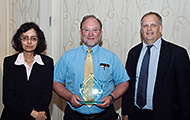 Photo of two men and one woman posing for a group picture. The man in the center is holding an award.