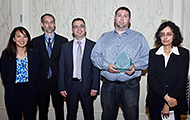 Photo of three men and two women posing for a group picture. One of the men in the center is holding an award.