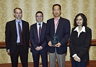 Photo of three men and one woman posing for a group picture. One of the men is holding an award.