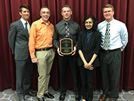 Photo of four men and one woman posing for a group picture. One man in the center is holding an award.