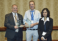 Photo of two men and one woman posing for a picture. Both men are holding an award.