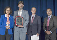 Photo of one woman and three men posing for a group picture. One man in the center is holding an award.