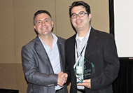 Photo of two men posing for a picture. The man on the right is holding an award.