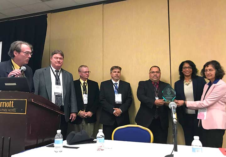 Pete Devlin and Sunita Satyapal present an award plaque to five staff from the United States Postal Service.