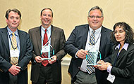 Photo of three men and one woman posing for a group picture. Two of the men are holding awards.